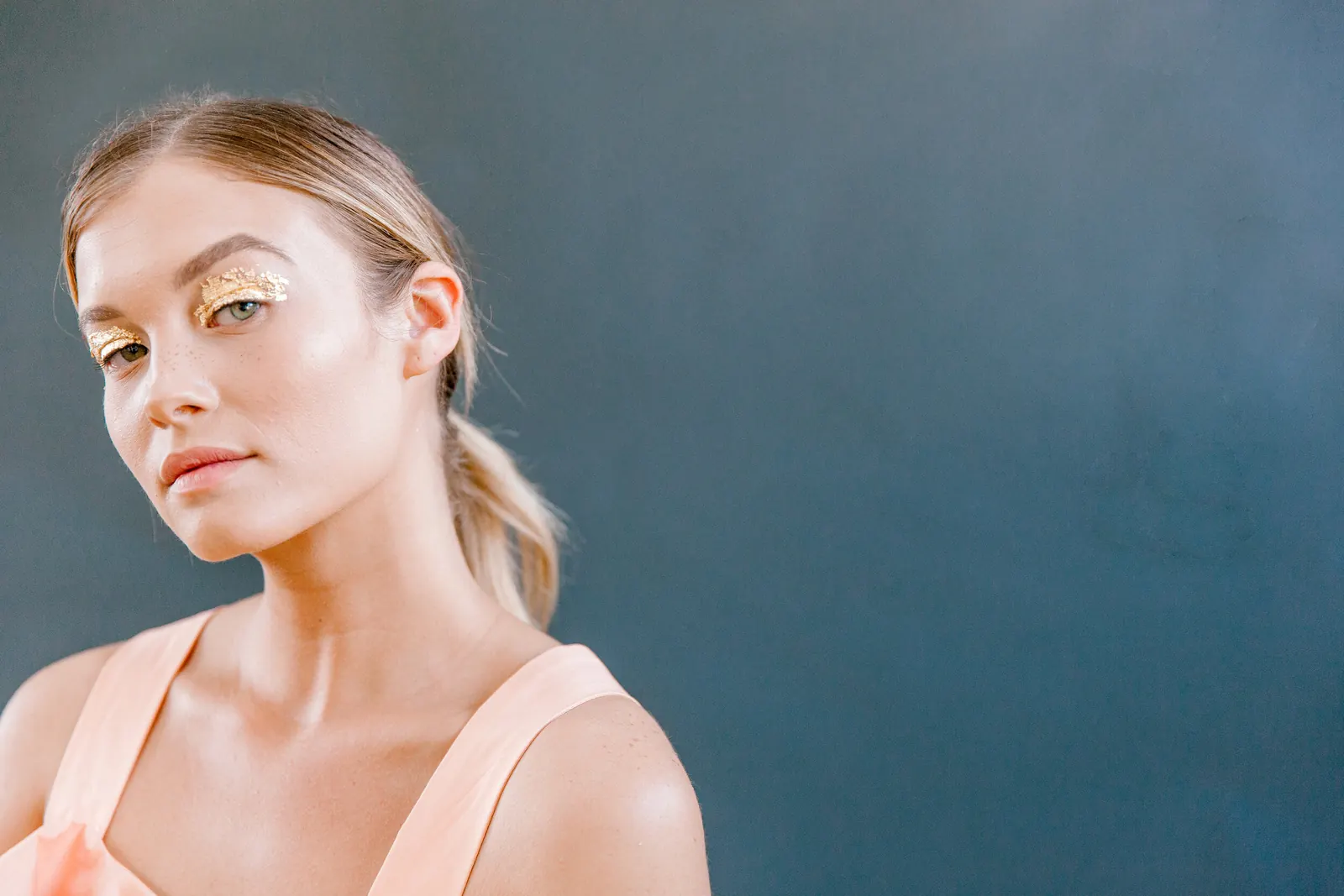 Woman with gold glitter eye makeup and soft peach lips against a blue backdrop
